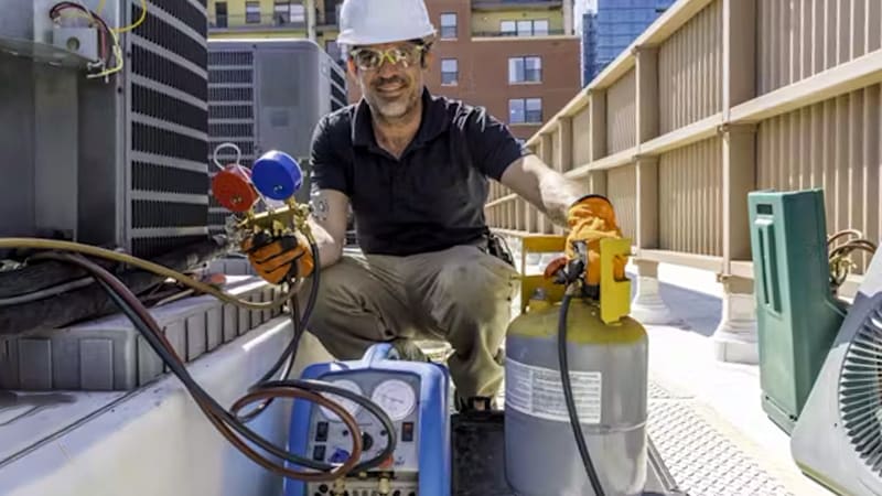 A HVAC technician properly outfitted with safety glasses and thick work gloves, carefully monitoring a recovery machine and tank