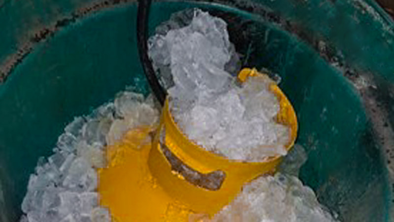 A refrigerant recovery tank sitting in a 5-gallon bucket filled with ice and water, with thick, large-diameter hoses connected to it