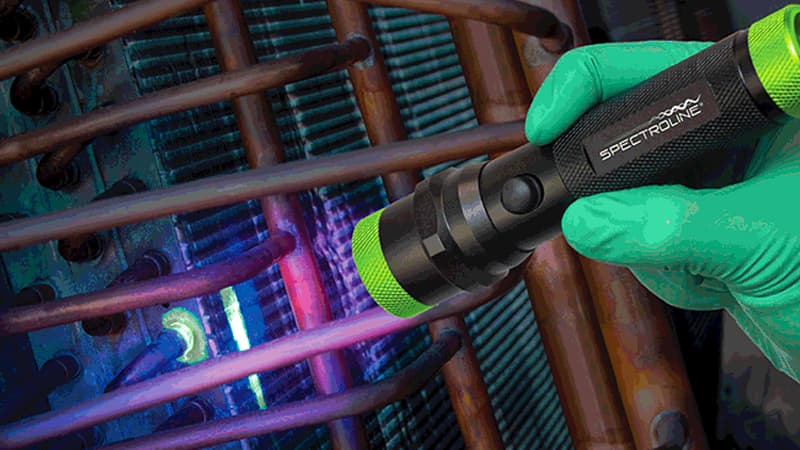 Technician using a UV flashlight to inspect HVAC coils for fluorescent dye indicating a leak