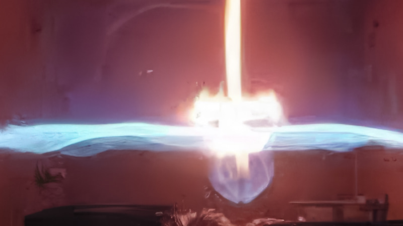 A controlled laboratory test showing a technician safely holding a lit match well above a small, weak flame originating from an A2L refrigerant source, demonstrating its low burning speed
